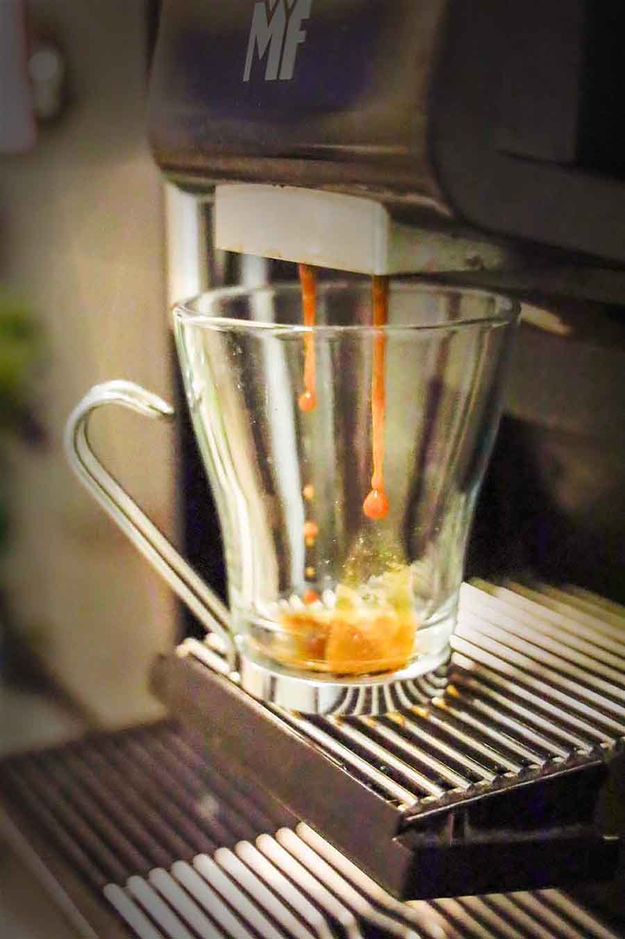 Eine Kaffeemaschine füllt frisch gebrühten Espresso in eine transparente Glastasse mit Henkel. Zwei kräftige, dunkelbraune Kaffeestrahlen fließen aus dem Auslauf der Maschine in die Tasse, die auf einem Abtropfgitter steht.