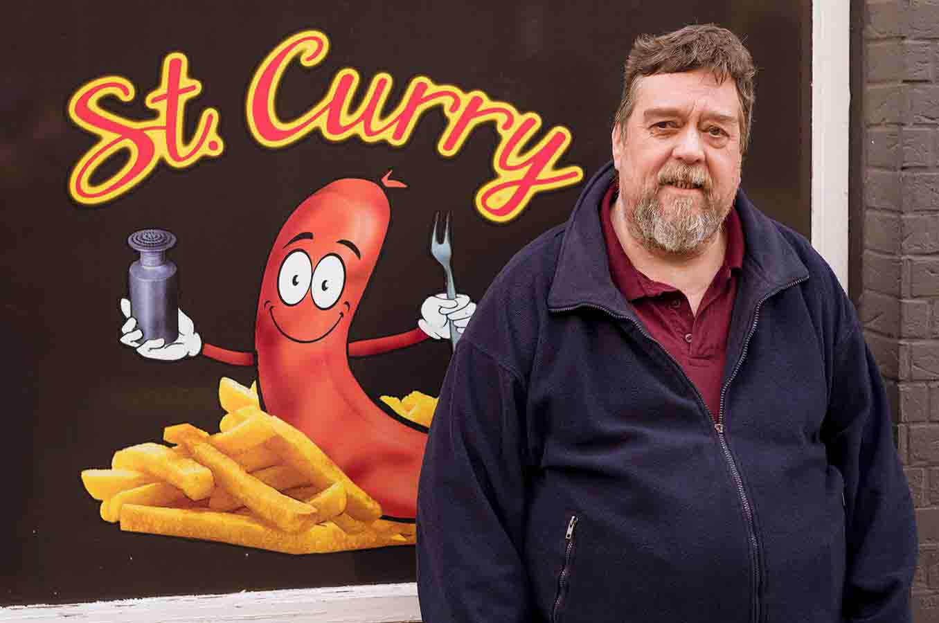 Ein bärtiger Mann steht vor einem Schaufenster mit der Aufschrift „St. Curry“ und einer Comicfigur in Form einer Wurst auf Pommes, die eine Gabel und einen Gewürzstreuer hält.