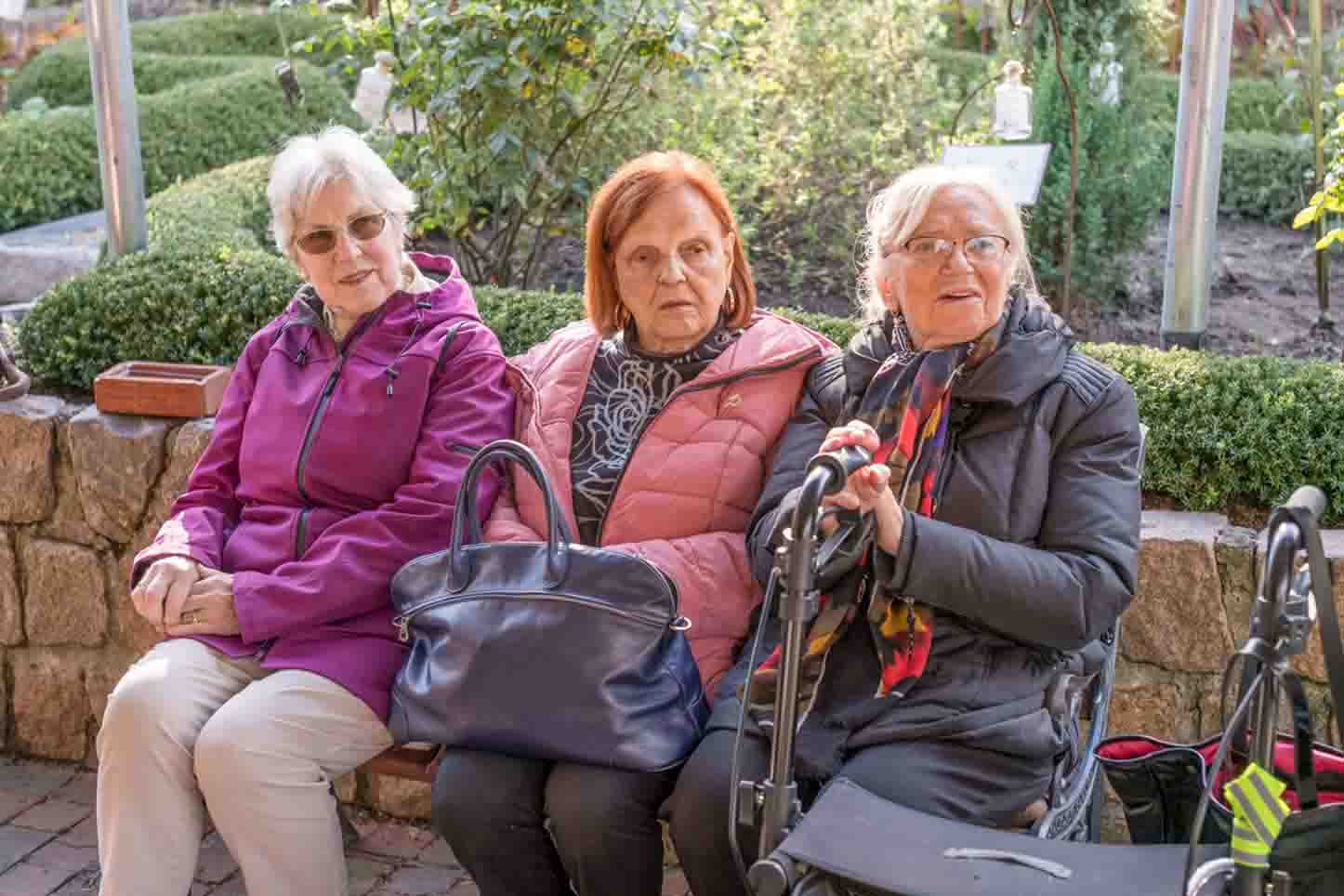 Drei ältere Frauen sitzen auf einer Bank im Freien und unterhalten sich. Sie tragen warme Jacken und haben Gehhilfen sowie Handtaschen bei sich. Im Hintergrund ist ein gepflegter Garten mit Hecken und Rosen zu sehen.
