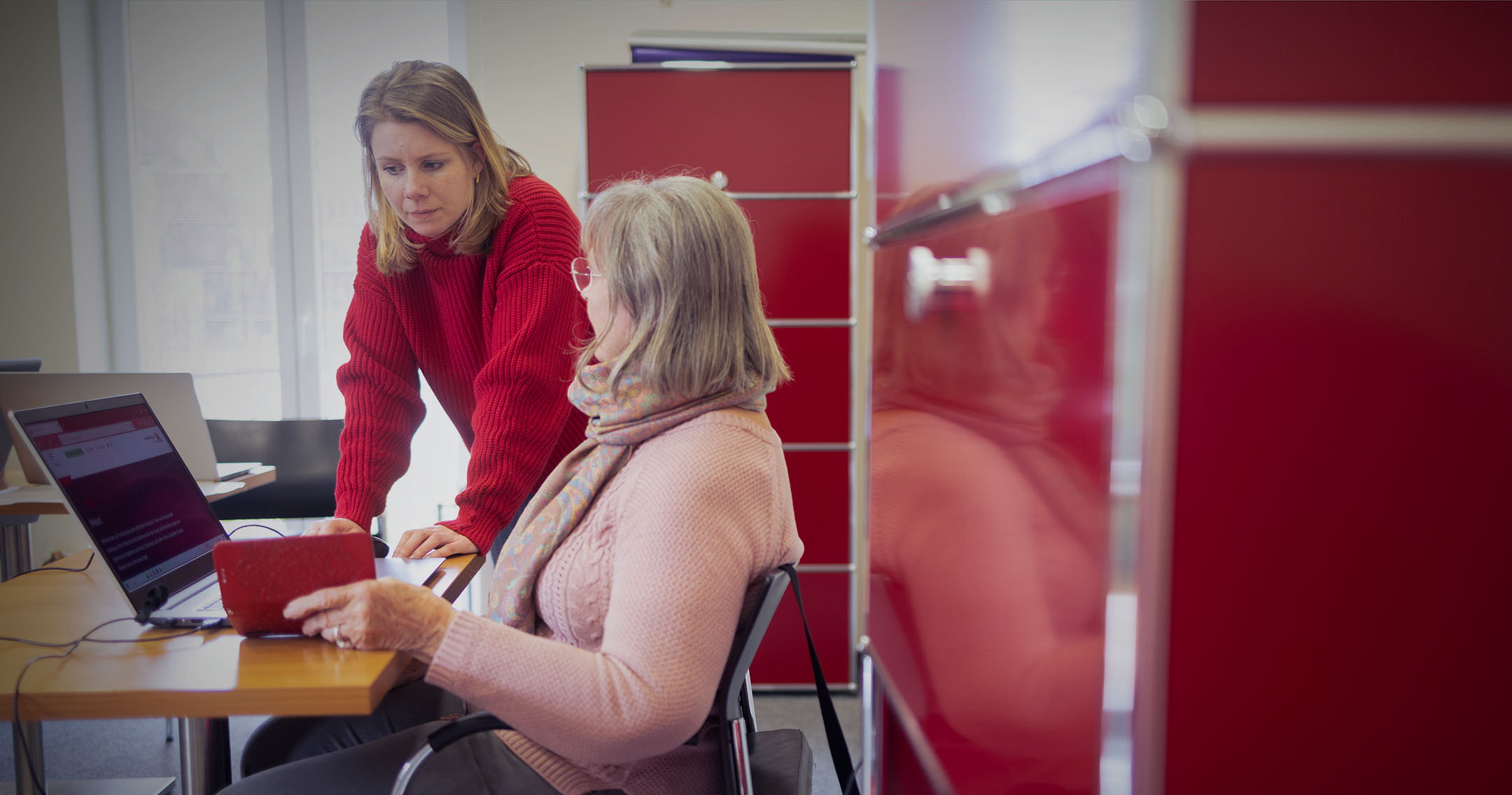 Eine jüngere Frau steht neben einer älteren Frau. Beide schauen auf einen Laptop auf einem Tisch in einem modernen Schulungsraum. Die Jüngere erklärt der Älteren etwas am Computer. Im Hintergrund sind weitere Laptops und rote Aktenschränke zu sehen.