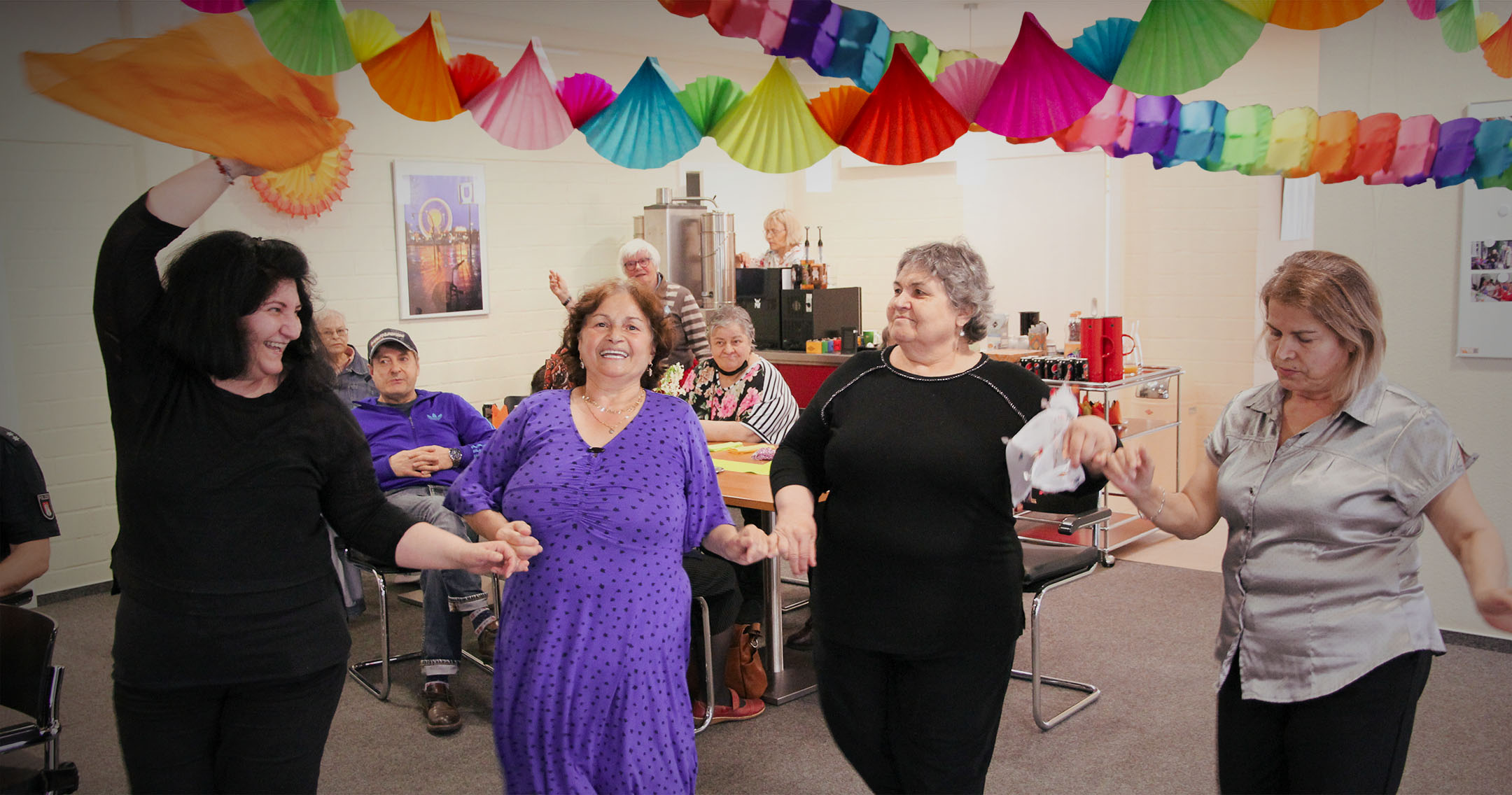 Eine Gruppe älterer Frauen tanzt fröhlich in einem festlich geschmückten Raum. Die Frauen halten sich an den Händen und lächeln, während sie bunte Tücher in der Luft schwingen. Im Hintergrund sitzen weitere Seniorinnen und Senioren an einem Tisch und schauen zu. Die Decke ist mit bunten Papiergirlanden dekoriert, was dem Raum eine festliche und fröhliche Atmosphäre verleiht.