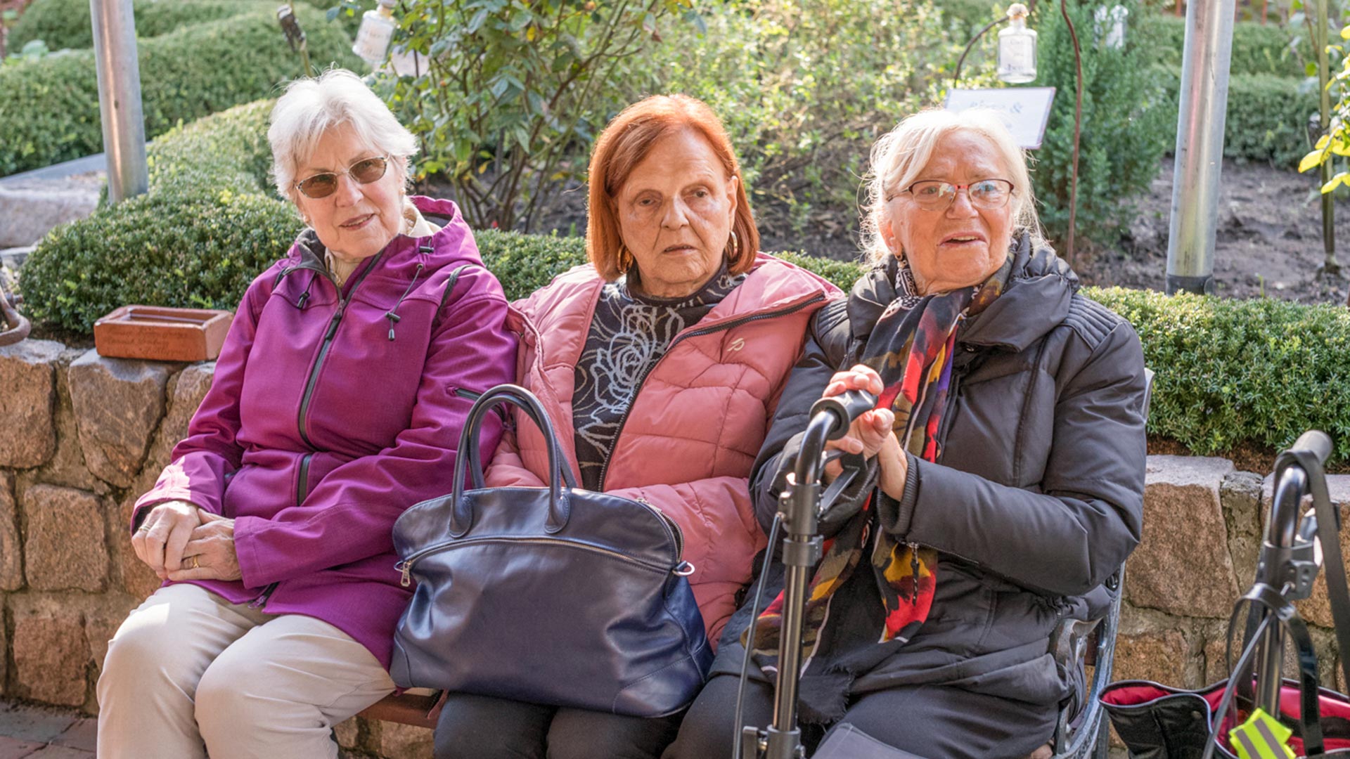 Drei ältere Frauen sitzen nebeneinander auf einer Bank im Freien und unterhalten sich. Sie tragen warme Jacken und haben Taschen oder Gehhilfen bei sich.