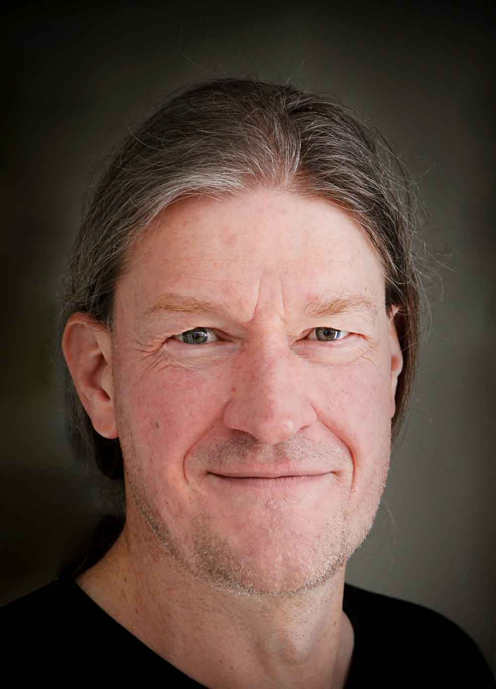 Porträt eines lächelnden Mannes mit langen grauen Haaren vor dunklem Hintergrund